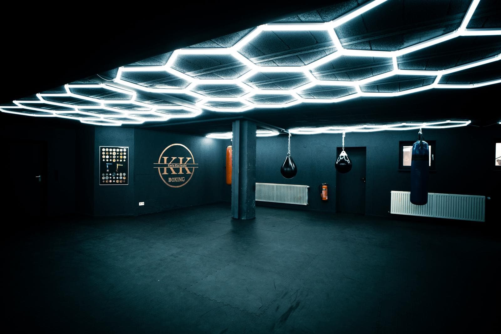 Die leere Halle von KK Boxing: Wabendecke aus weißen LED-Röhren, KK-Logo und ein Rahmen mit Medaillen an der Wand, Säcke an Ketten, Mattenboden